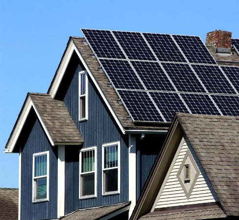 A house with dark blue wooden siding and a gabled roof is equipped with solar panels. The sky is clear and blue.
