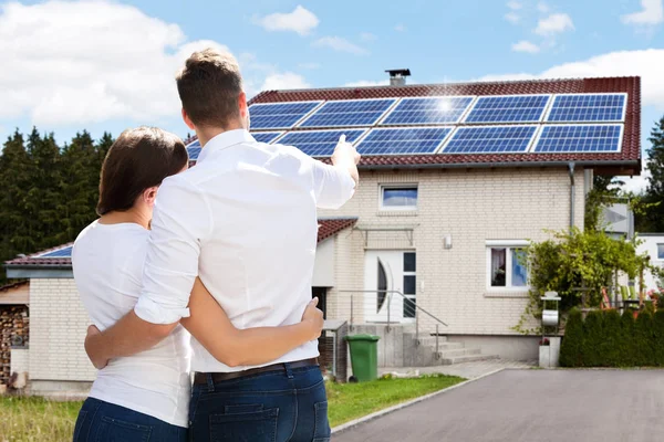 A couple stands in front of a white house with solar panels on the roof. The man points towards the solar panels as they embrace. The house is set in a suburban area with trees in the background.