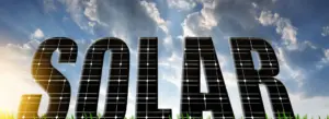 The word "SOLAR" appears in large, capital letters with a texture resembling solar panels. The letters are positioned against a backdrop of a partly cloudy sky and tall green grass at the bottom. Sunlight is visible, creating a bright and clear atmosphere.