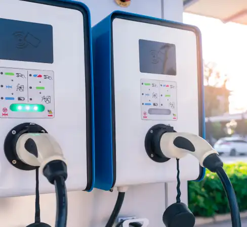 Two electric vehicle charging stations with cables and plugs attached. The units display indicator lights and operational instructions. A blurred background shows greenery and soft daylight.