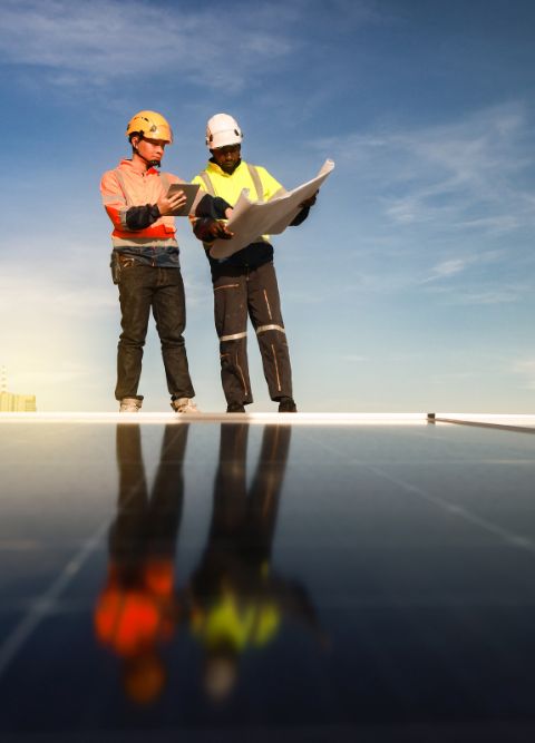 Two construction workers wearing helmets and safety vests are standing on a rooftop with solar panels, discussing a blueprint. The blue sky is visible in the background. One worker points at the blueprint while the other holds it open.