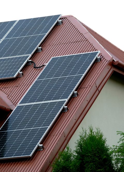 Solar panels are mounted on a red, corrugated metal roof of a house. The panels are angled to capture maximum sunlight. Green foliage from nearby trees is visible in the foreground. The sky is overcast, providing a neutral backdrop to the setup.