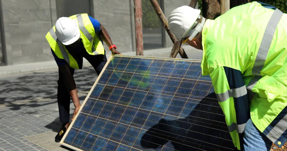 Two workers wearing safety vests and hard hats are positioning a solar panel outdoors. One worker is lifting the panel while the other supports it. They are in a paved area with a building in the background.