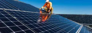 A worker in a high-visibility orange jacket and white hard hat is crouched on a rooftop, engaged in the solar panel installation process under a bright blue sky. The shiny panels, forming a grid-like pattern across the roof, reflect the precision embodied by New Yorker Solar.